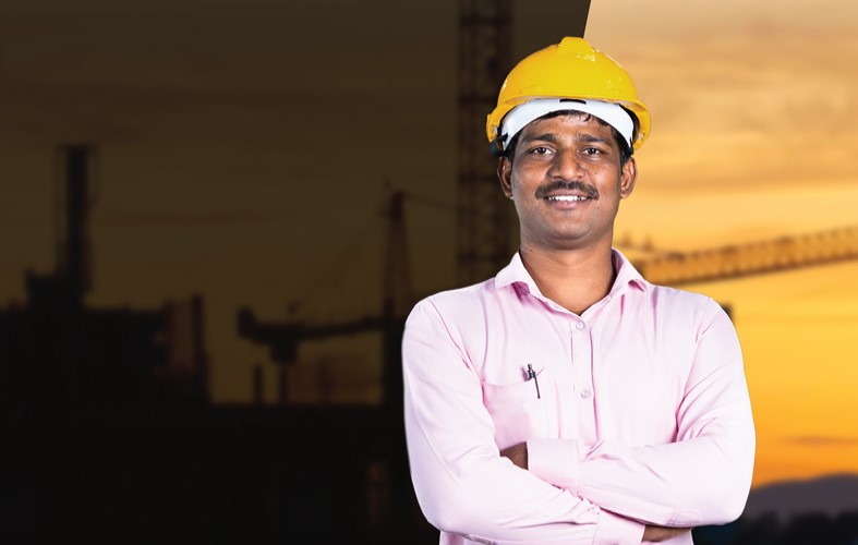 A man wearing a yellow hard hat and pink shirt, arms crossed, stands in front of a backdrop showing a construction site at sunset. The left side is shadowed, contrasting with the bright, orange sky on the right.
