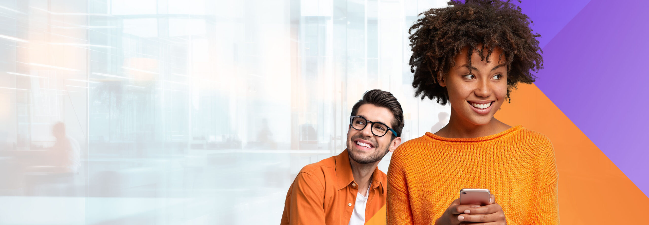 A woman with curly hair is smiling and holding a phone, wearing an orange sweater. A man with glasses and a beard, wearing an orange shirt, is smiling and looking up. The background is a blurred modern office with orange and purple diagonal lines.