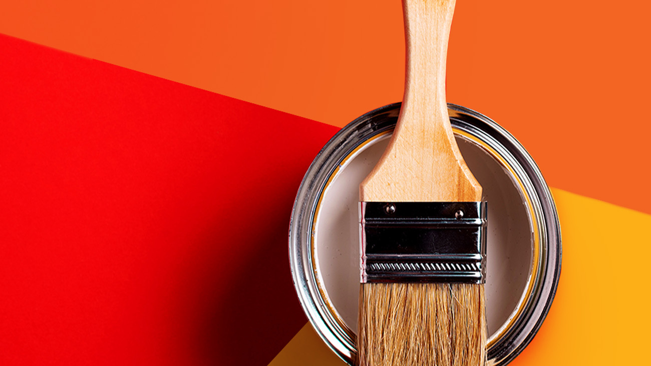 A wooden paintbrush rests on an open can of white paint against a vibrant background of red, orange, and yellow geometric shapes.