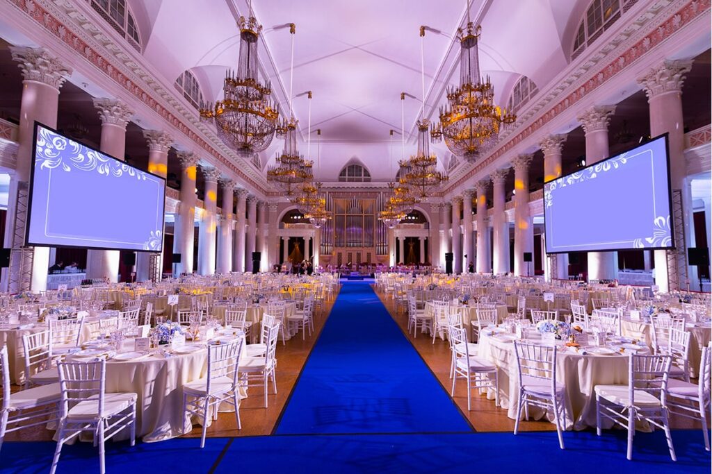 An elegant ballroom set for an event, featuring a long blue carpet leading to a stage. Round tables with white chairs fill the room, adorned with floral centerpieces. Large chandeliers hang from the high ceiling, and two big screens display ornamental designs.