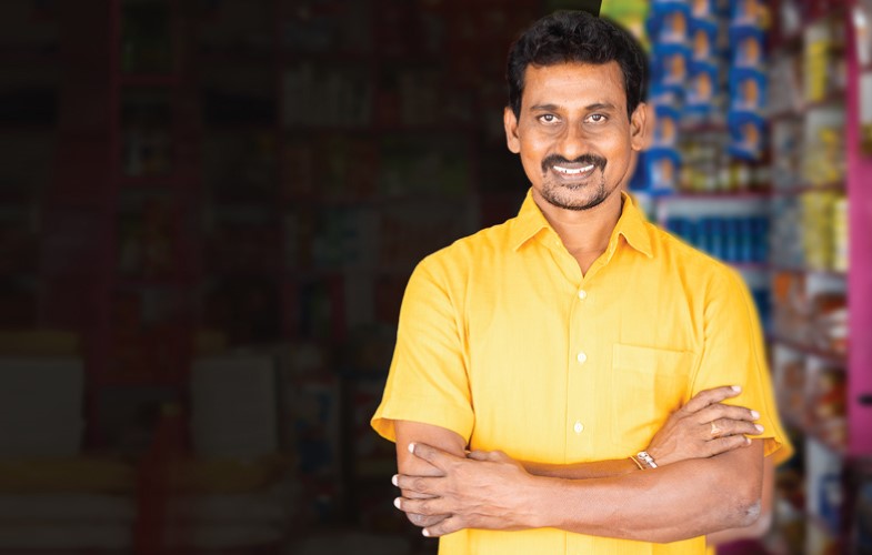 A man in a bright yellow shirt smiles with arms crossed in front of a store background. The store shelves behind him are filled with colorful products, slightly out of focus.
