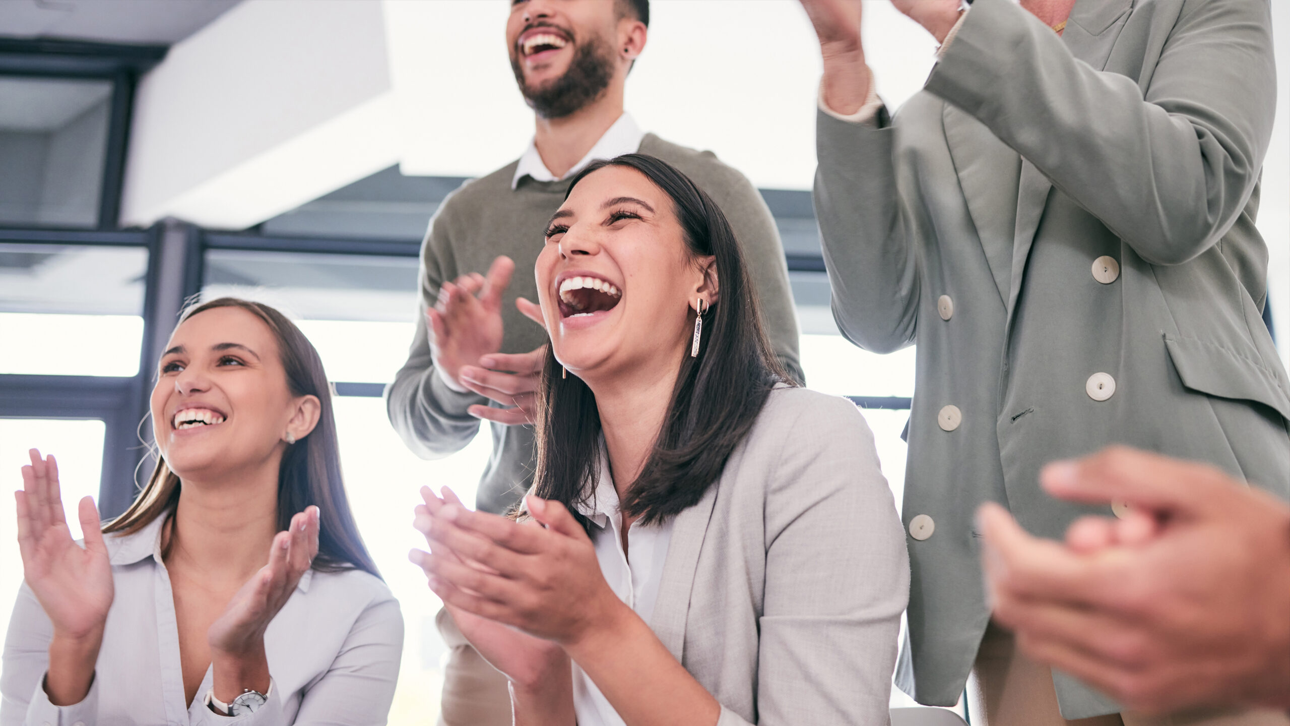 A group of people are standing and clapping, with big smiles on their faces. They appear to be in a bright, modern office setting. The mood is joyful and celebratory.