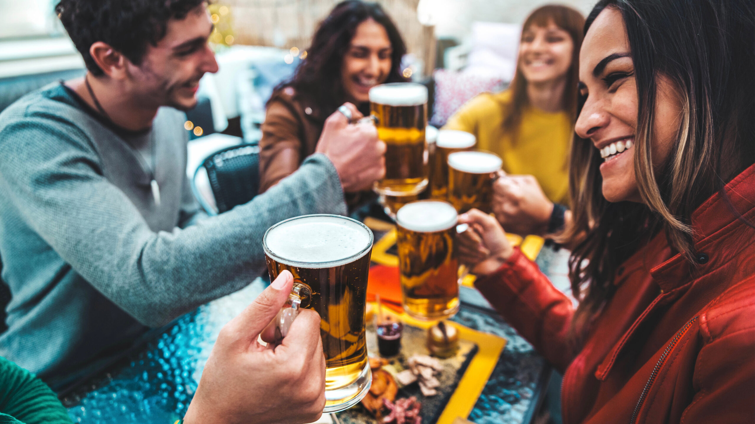 A group of five people sit around a table, smiling and raising glasses of beer in a toast. The table has small dishes of snacks. The scene is lively and joyful, suggesting a social gathering.