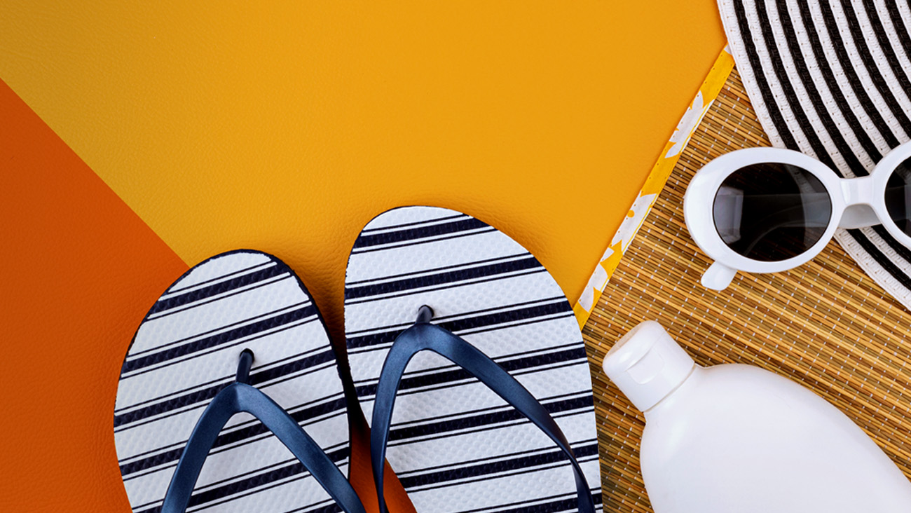 A flat lay image featuring black and white striped flip-flops, white sunglasses, a round bottle of sunscreen, and a striped sun hat on an orange surface, evoking a summer or beach theme.