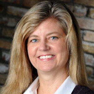 A woman with long blonde hair and blue eyes, smiling and wearing a white blouse and dark blazer, stands against a blurred brick wall background.