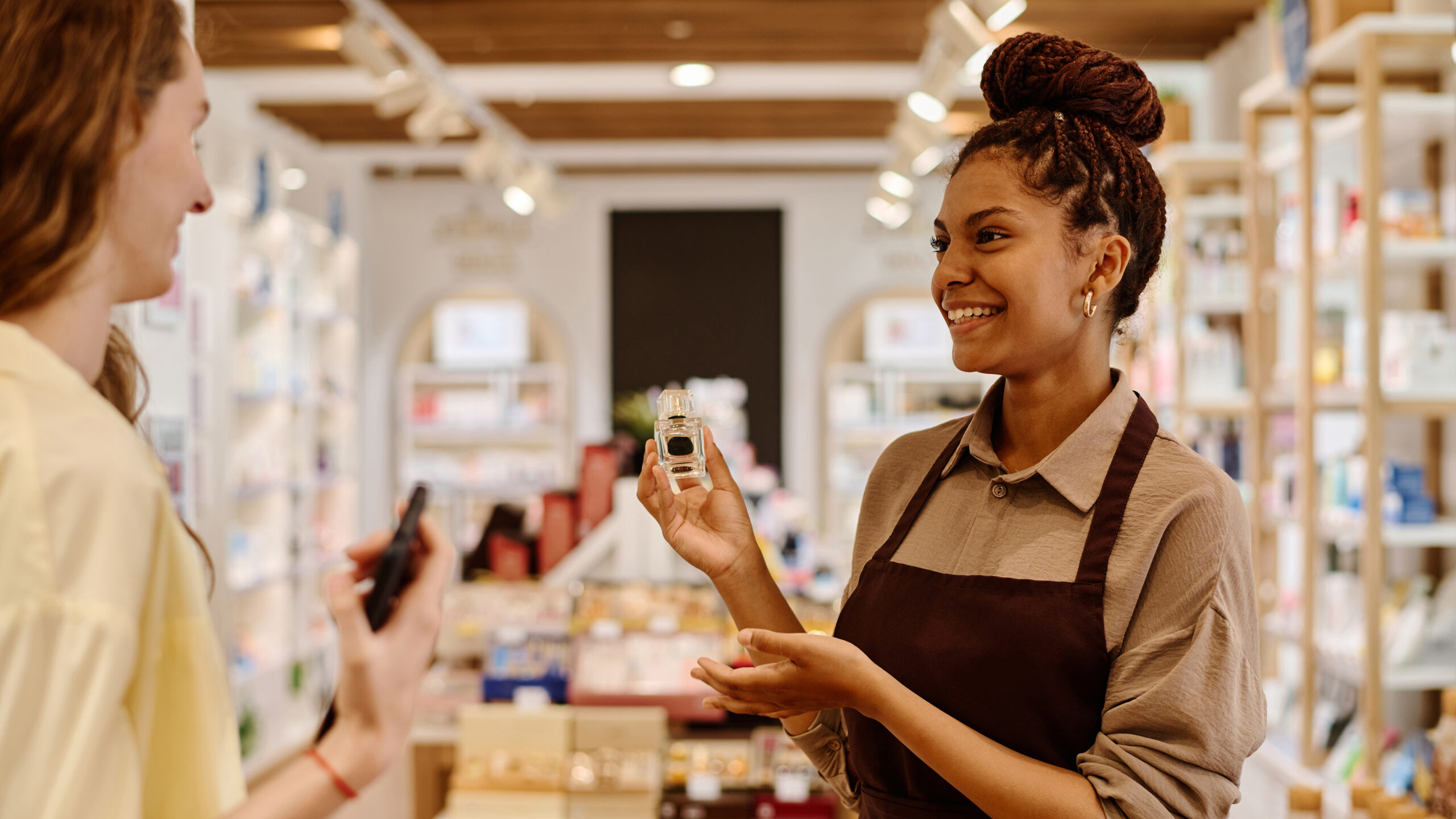 A store employee wearing a brown apron shows a bottle of essential oil to a customer. They are smiling at each other in a well-lit shop filled with shelves of various products.