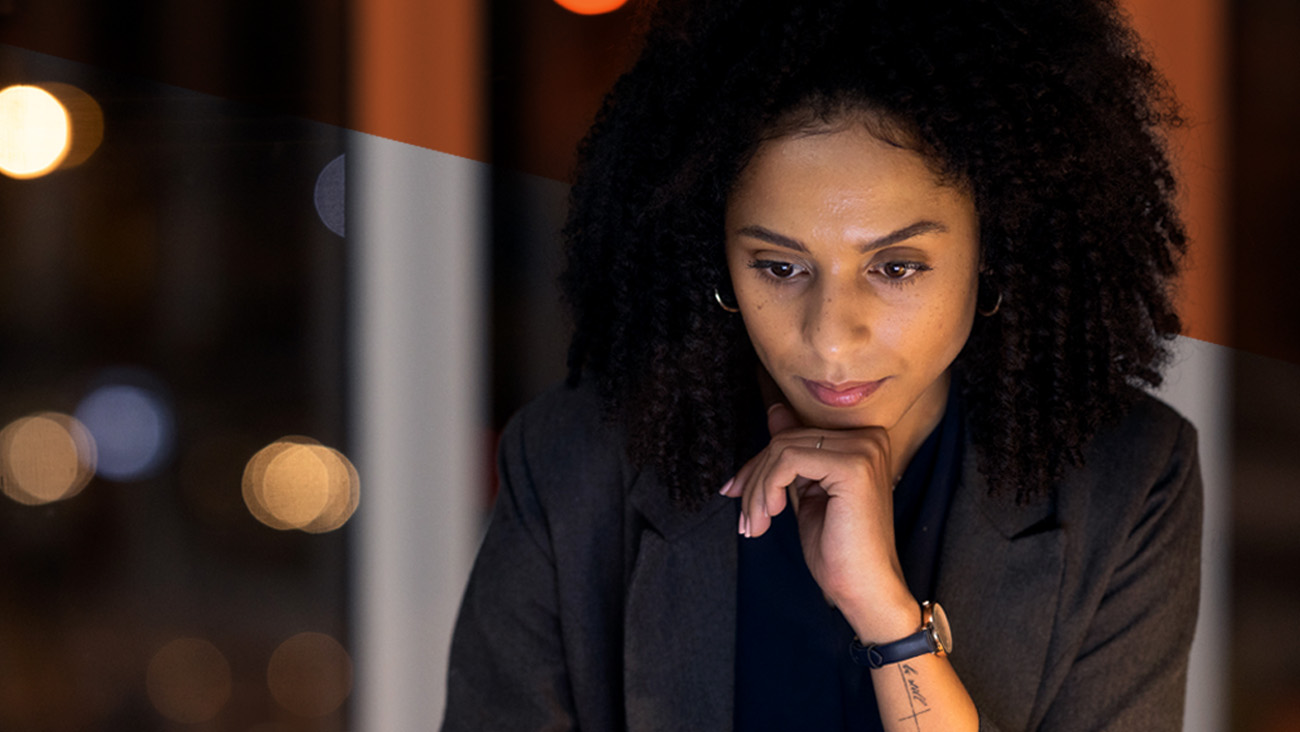 A person with curly hair, wearing a dark blazer, appears deep in thought while working on a laptop. The background shows out-of-focus, warm-toned lights, suggesting an evening or nighttime setting.