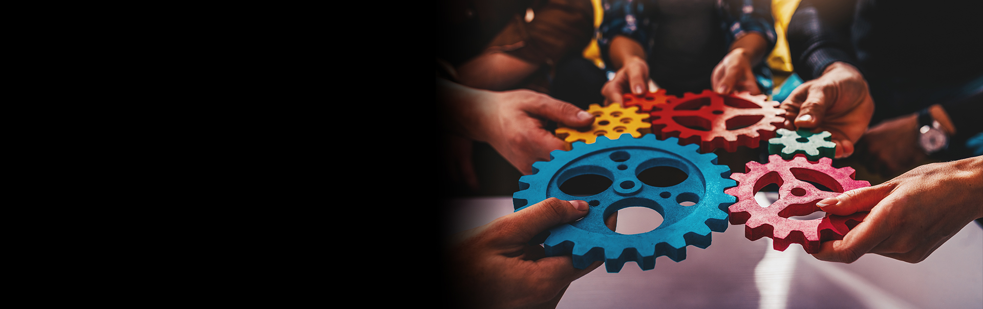 Several hands are holding colorful, interlocking gears of different sizes, symbolizing teamwork and collaboration. The background is slightly blurred, with a focus on the gears in red, blue, yellow, and orange. The left side is dark.