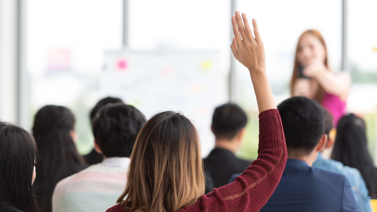 A woman raises her hand in a classroom setting, seen from behind. She is wearing a red sweater. In front is a blurry instructor speaking to the group, with a whiteboard and windows in the background.