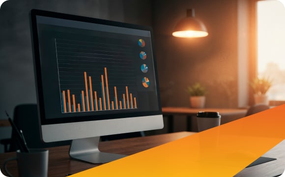 A computer screen displays a bar chart and pie charts with orange elements in a dimly lit office. A round light hangs from the ceiling, and a cup sits on the desk. Sunlight streams through a window, adding a warm glow to the room.