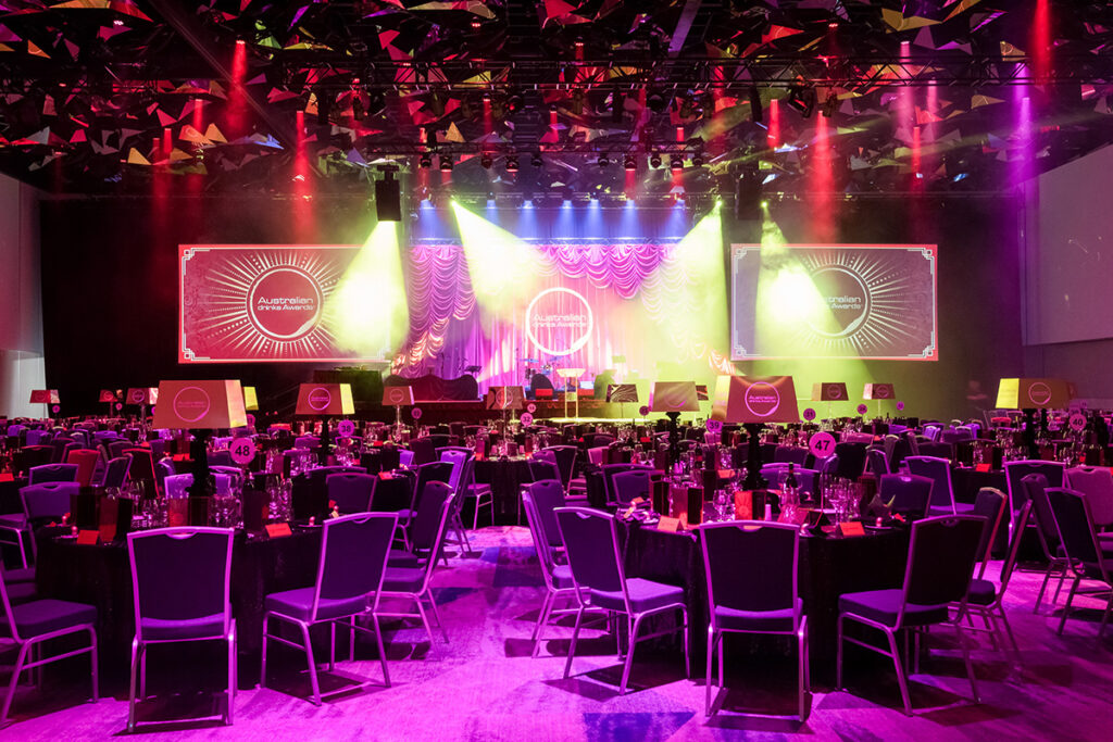 A large banquet hall with purple-lit tables and chairs is set up for an event. A stage with red curtains and colorful lights is at the front, displaying circular logos and text. Spotlights create a vibrant atmosphere.