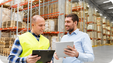 Two men are standing in a warehouse. One wears a yellow safety vest and holds a clipboard, while the other is in a light blue shirt, holding a tablet. They are smiling and appear to be discussing something. Shelves stacked with boxes are in the background.
