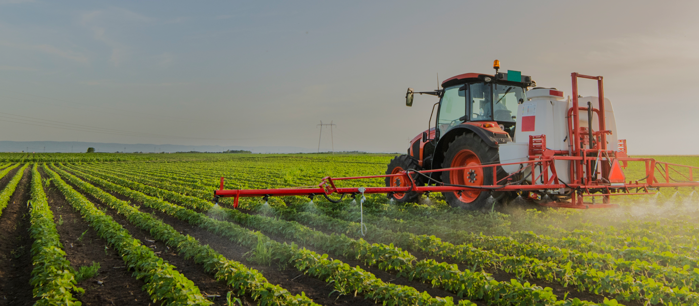 A red tractor sprays crops with pesticides in a sprawling green field. The tractor moves through neatly arranged rows of plants, with a clear sky and distant mountains in the background.