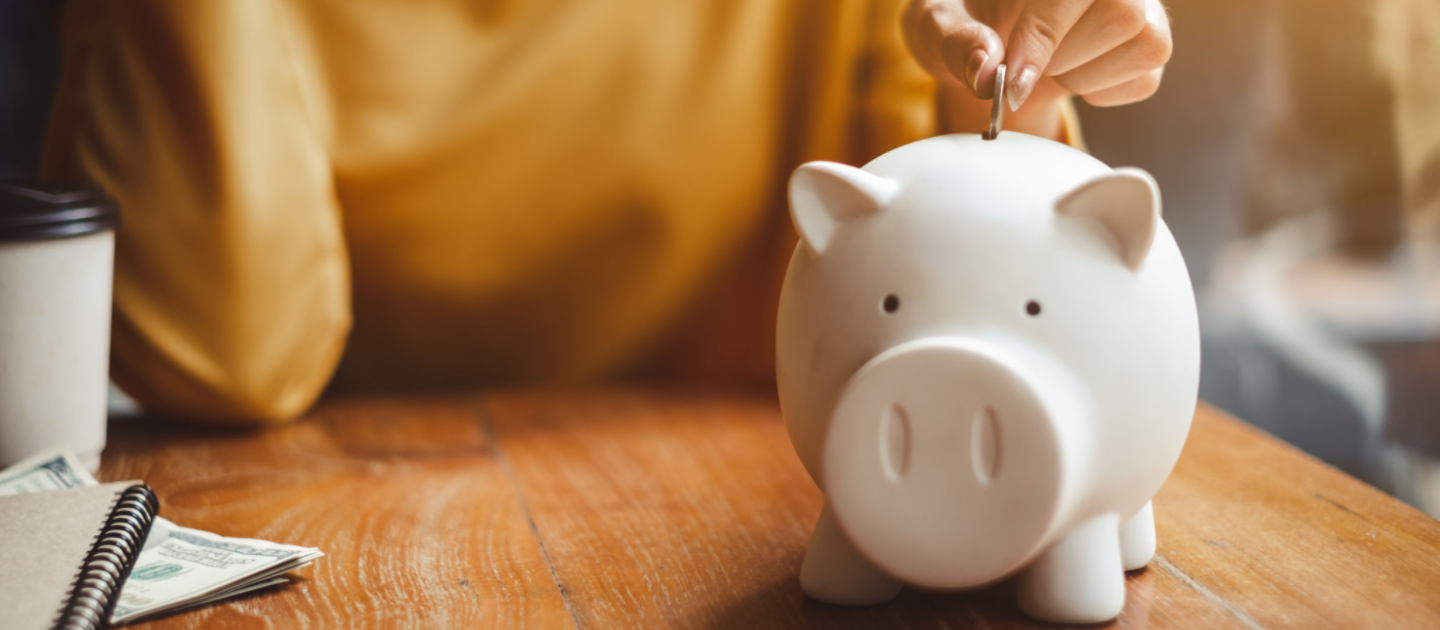 A person in a yellow shirt places a coin into a white piggy bank on a wooden table. A notebook, dollar bills, and a takeaway coffee cup are nearby, suggesting saving or financial planning.