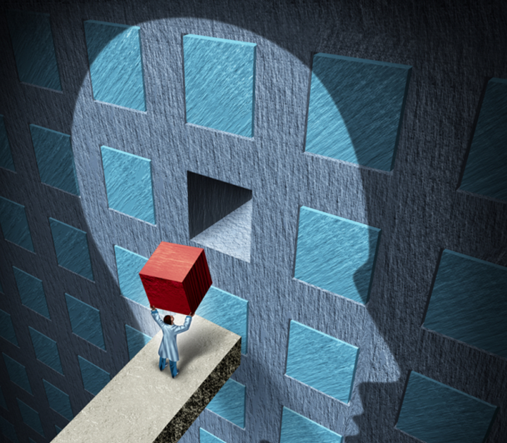 A person in a lab coat stands on a narrow pathway, lifting a red cube towards a human head silhouette formed by missing blue square tiles on a large wall. The cube fits into a matching space in the head silhouette, symbolizing fitting ideas or solutions.
