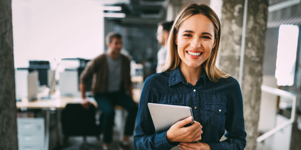 A woman with long hair and a denim shirt smiles while holding a tablet. She stands in a modern office setting, with blurred coworkers and desks in the background.