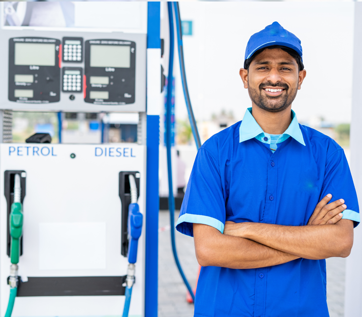 A man in a blue uniform and cap stands confidently with arms crossed next to a fuel pump at a gas station. The pump has green and black hoses labeled Petrol and Diesel. The background is slightly blurred.