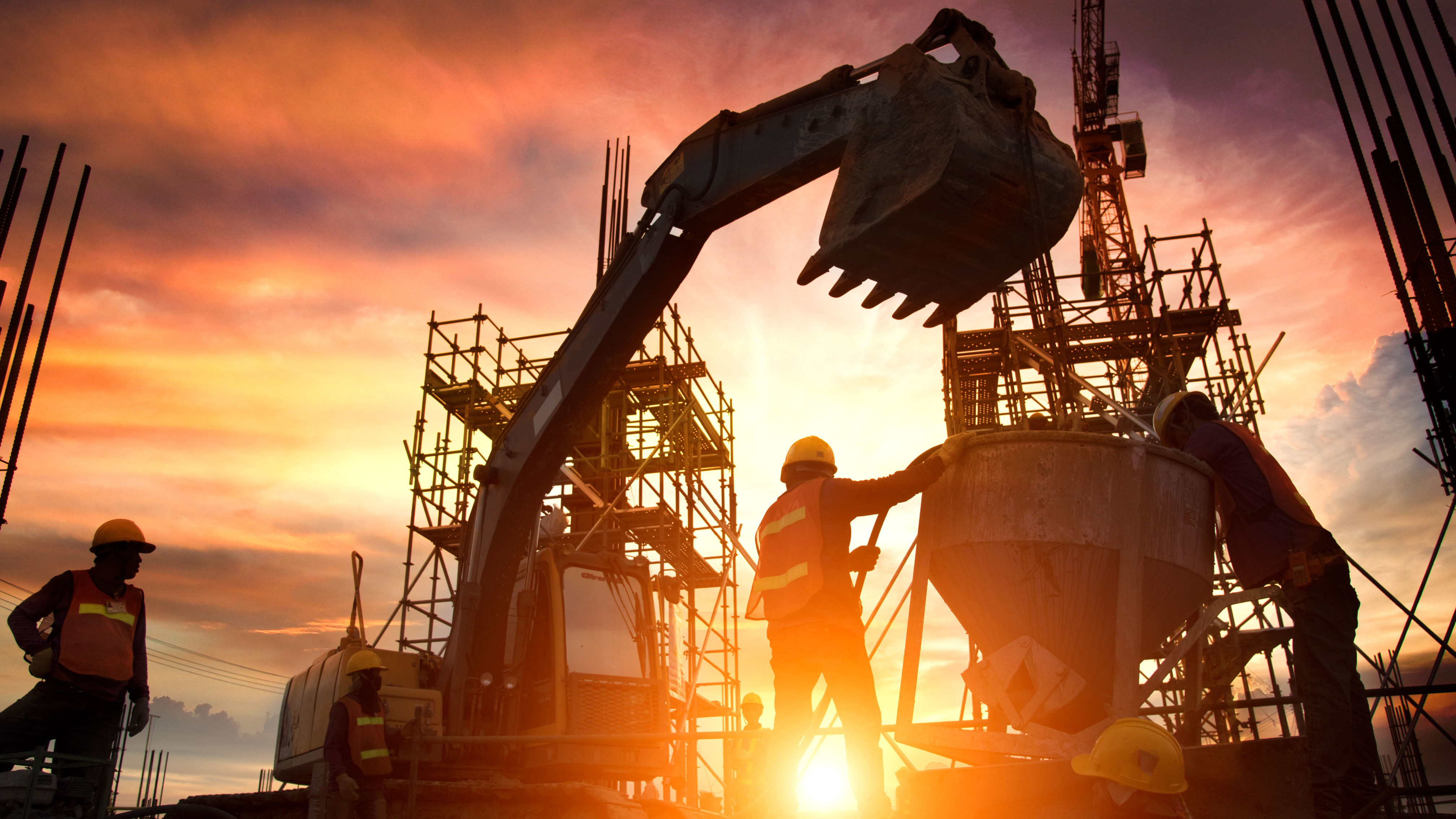 Silhouetted construction workers and an excavator are active on a site during a dramatic sunset. Metal scaffolding and cranes tower in the background, with the sun glowing brightly at the horizon, casting warm colors across the sky.