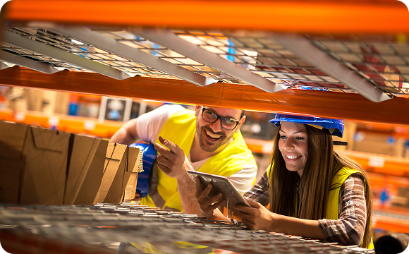 Smiling man and woman in safety vests and helmets use a tablet in a warehouse. They crouch by metal shelving with cardboard boxes in the foreground, suggesting collaboration and inventory management.