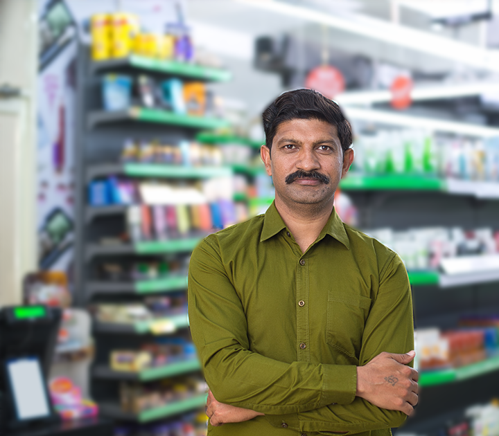 A man with a mustache, wearing a green shirt, stands with crossed arms in a store aisle. Shelves stocked with various colorful products are blurred in the background.