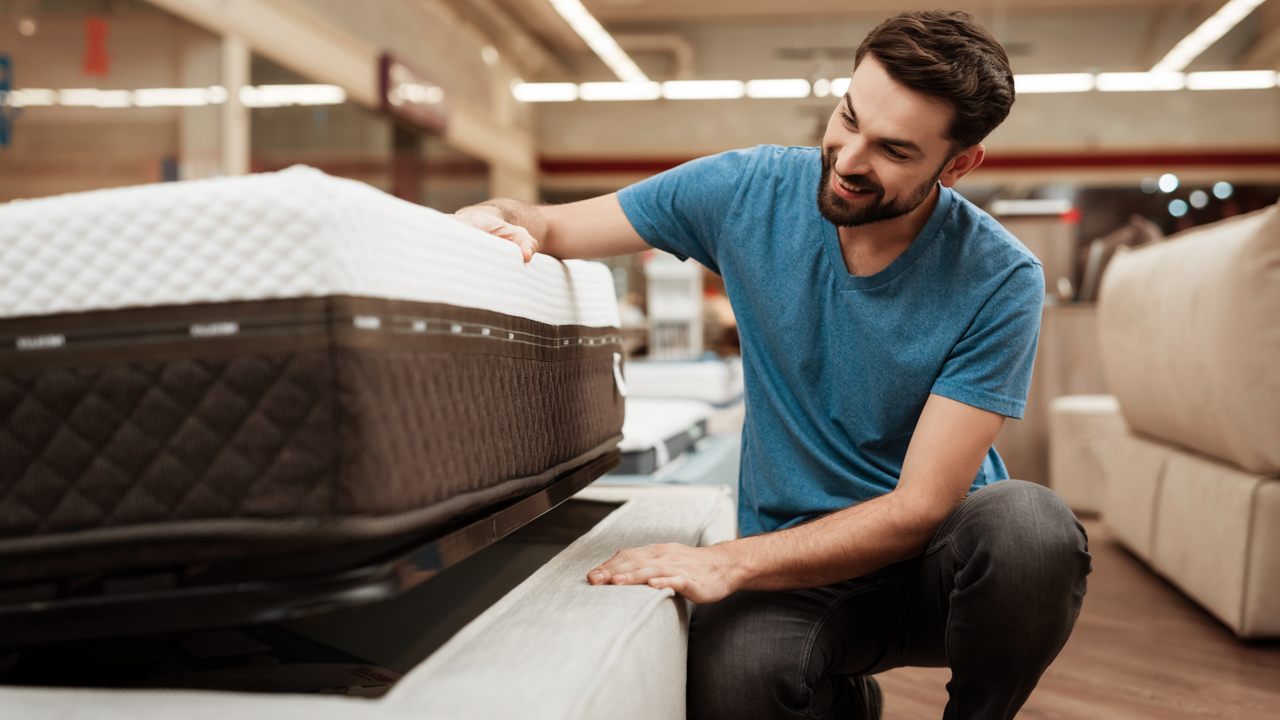 A man in a blue t-shirt is examining a mattress in a store. He is touching the corner of the mattress while kneeling, with various other mattresses in the background. The store has bright lighting and a modern interior.
