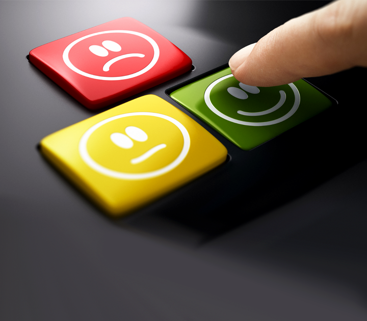 A finger presses a green button with a smiling face on a device with three buttons. The other buttons are yellow with a neutral face and red with a sad face, suggesting a system for feedback or rating.