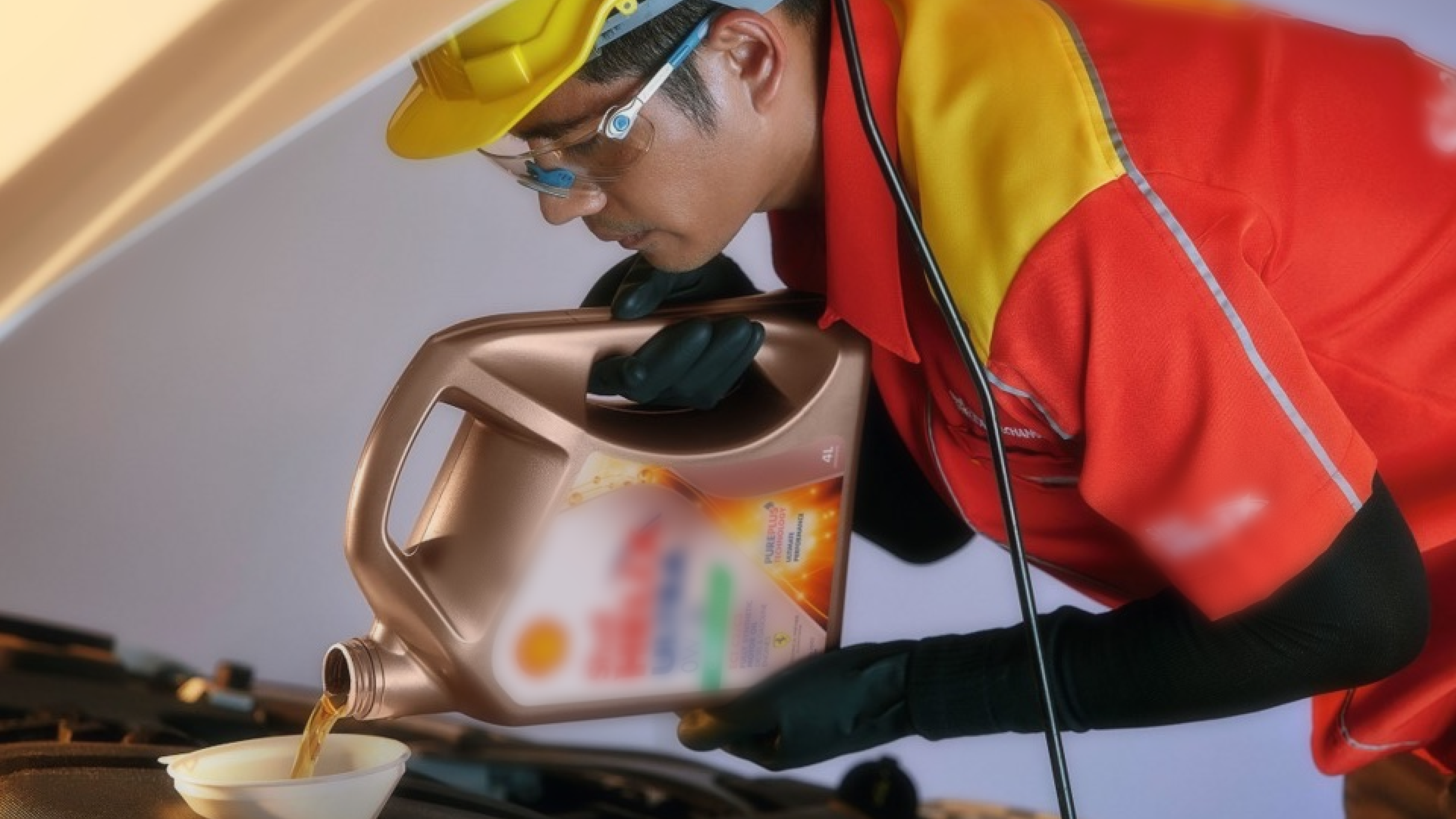 A worker in a red and yellow uniform and safety gear pours motor oil from a large container into a funnel placed under a car hood. The worker wears gloves, a hard hat, and goggles, focusing on the task.