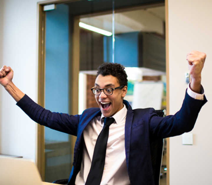 A person in a suit and tie sits at a desk, raising their fists in excitement, with a wide smile. They are in a modern office space with glass walls.
