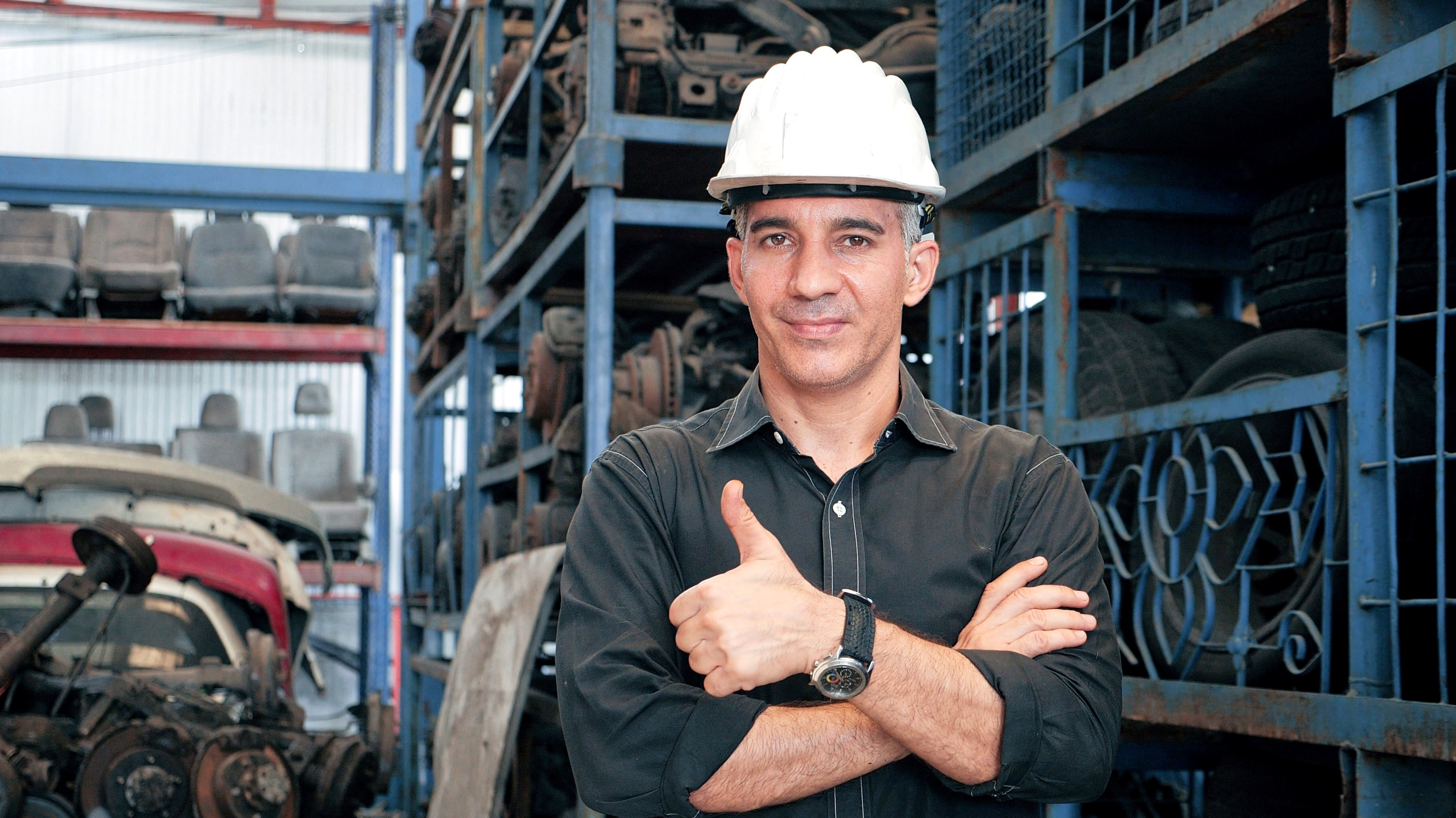 A man in a black shirt and white hard hat stands in a warehouse filled with car parts, giving a thumbs-up with one hand while the other is crossed over his chest.